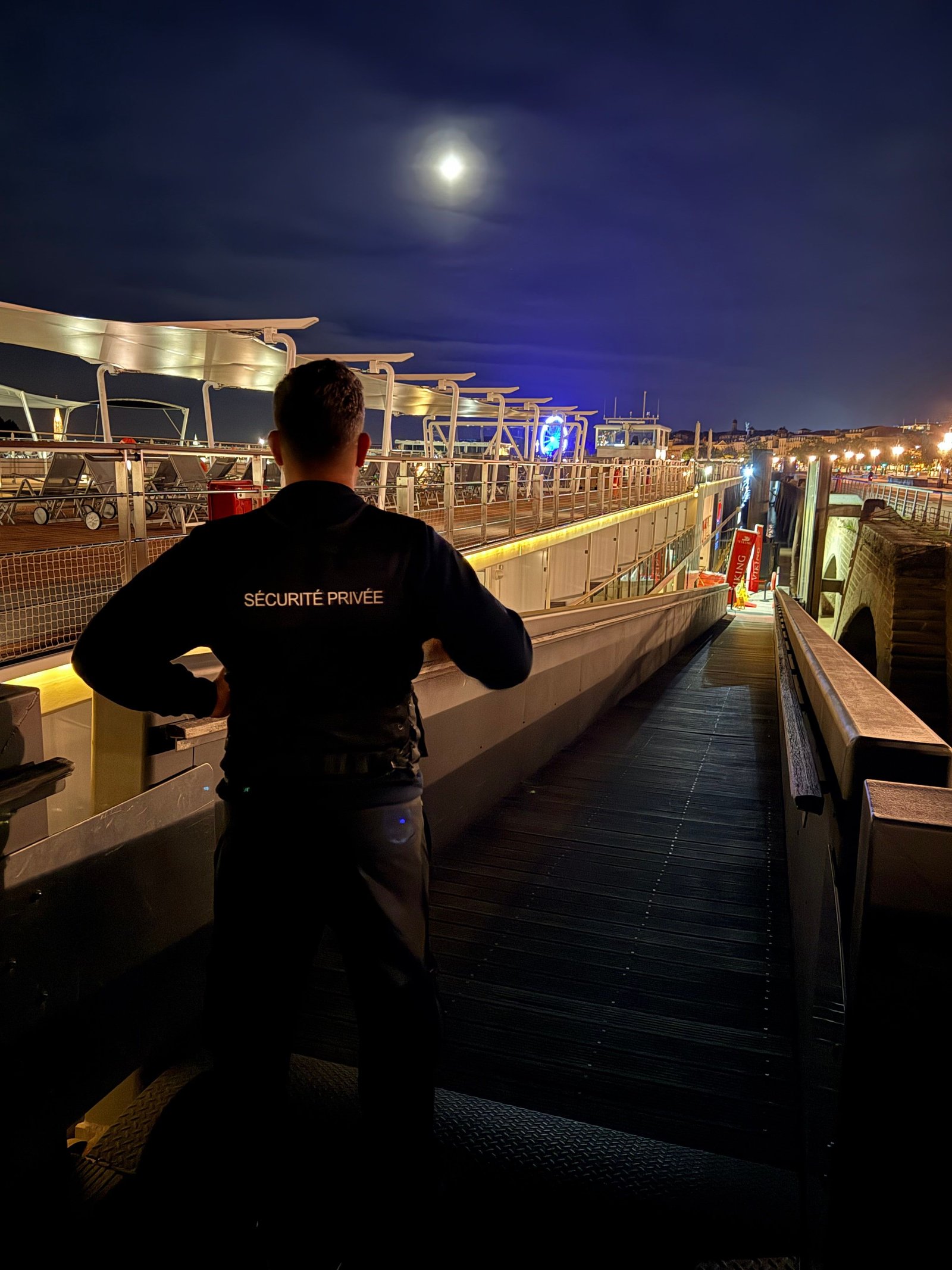Agent de sécurité de dos, de nuit, à l’accès d’un embarcadère sur les quais de Bordeaux